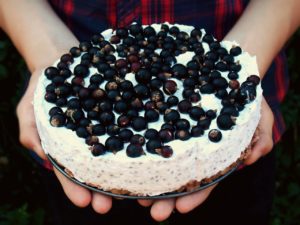 Ein Sommerkuchen mit dicker Cremeschicht und Beeren oben drauf wird von zwei Händen vorsichtig gehalten.