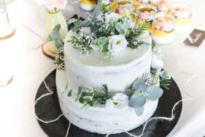 Auf einem schwarzen Teller steht ein zweistöckiger Hochzeitskuchen, der mit verschiedenen blauen und grünen Blumen und Blättern dekoriert ist.