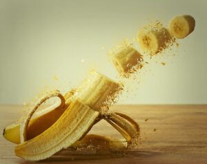 Eine Banane ist halb geschält und liegt auf einer Holzoberfläche. Sie ist in mehrere Teile geschnitten, welche stückweise wegfliegen.