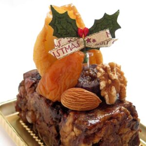 Auf einem kleinen Früchtebrot sind getrocknete Aprikosen, Nüsse und eine weihnachtliche Schleife drapiert.