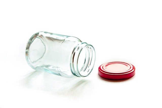 Ein sauberes, offenes Schraubglas liegt auf einem weißen Untergrund, ein roter Deckel daneben