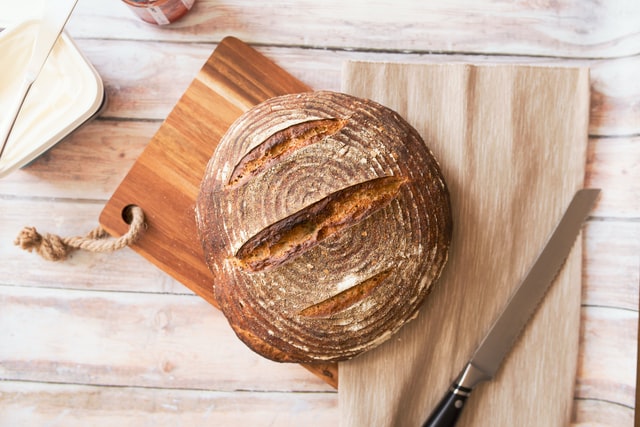 Auf einem Holzbrett liegt ein frisches Brot mit Einschnitten, daneben ein geriffeltes Brotmesser