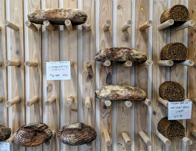 An einer mit Holz verkleideten Wand liegen verschiedene Brotlaibe auf Regalen aus Holzstäben zum Verkauf