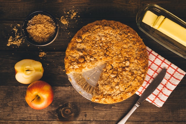 Auf einem dunklen Holztisch steht ein angeschnittener Apfelkuchen, daneben stehen verschiedene Zutaten
