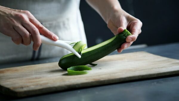Jemand schneidet eine Zucchini mit einem Sparschäler in dünne Längsstreifen