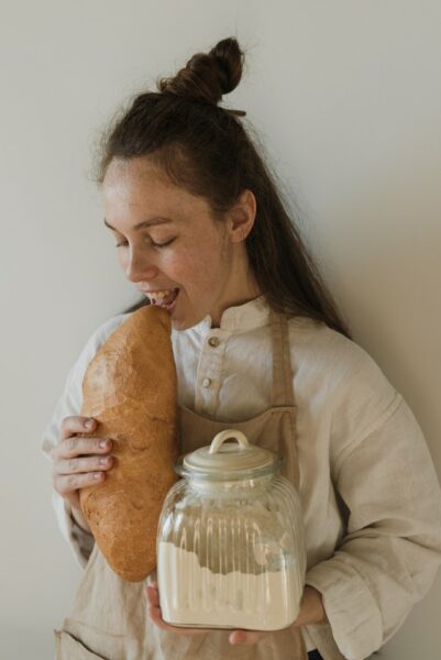 Eine junge Frau hält ein Vorratsglas mit Mehl im einen Arm und im anderen ein Brot, in dass sie beißt