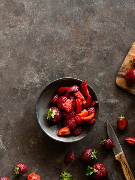 In einer kleinen Schale liegen mehrere geschnittene Erdbeeren, daneben ein Messer und weitere Früchte