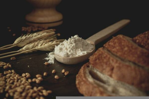 Neben einem in Scheiben geschnittenem Brot liegen ein Holzlöffel voll Mehl, einige Getreideähren und Körner