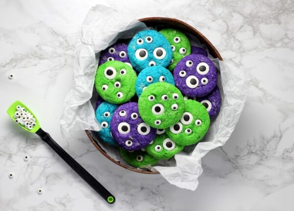 In einer Schüssel auf einem Tisch liegen grün, lila und blau gefärbte Kekse mit Deko-Augen 
