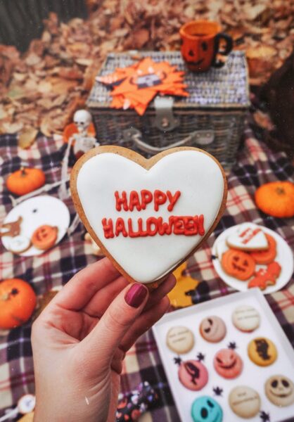 Eine Person hält einen herzförmigen Keks mit der Aufschrift "Happy Halloween", im Hintergrund sind mehr Kekse und Kürbisse