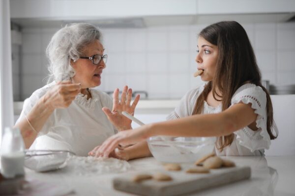 Eine ältere Dame erklärt etwas einem jungen Mädchen mit Keks im Mund. Vor Ihnen liegen Schüsseln und Mehl auf einem Tisch