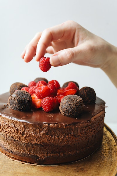 Eine Hand platziert mittig auf einen Schokoladenkuchen zu verschiedenen anderen Beeren noch eine weitere Himbeere
