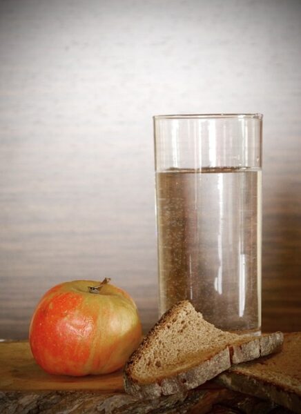 Neben einem gefüllten Wasserglas liegt eine trockene halbe Scheibe Brot und ein schon etwas verschrumpelter Apfel