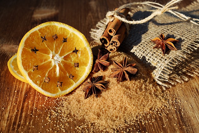 Auf einem Holztisch liegt eine mit Nelken gespickte Orangenscheibe, daneben etwas Jute, ein Bündel Zimt, ein Haufen brauner Zucker und einige Sternanis-Früchte