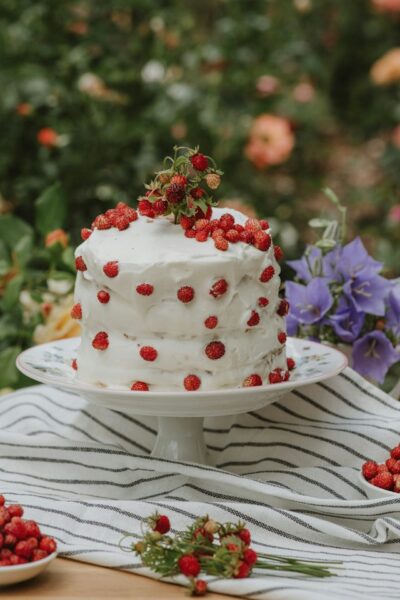 Auf einer einstöckigen Etagere steht ein hoher Kuchen, der mit einer weißen Creme überzogen und mit kleinen Walderdbeeren dekoriert ist.