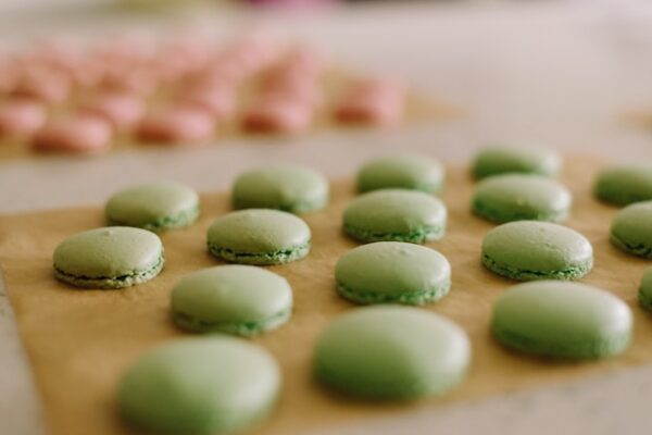 fertig gebackene grüne und rosafarbene Macaron-Hälften liegen aufgereiht auf Backpapieren